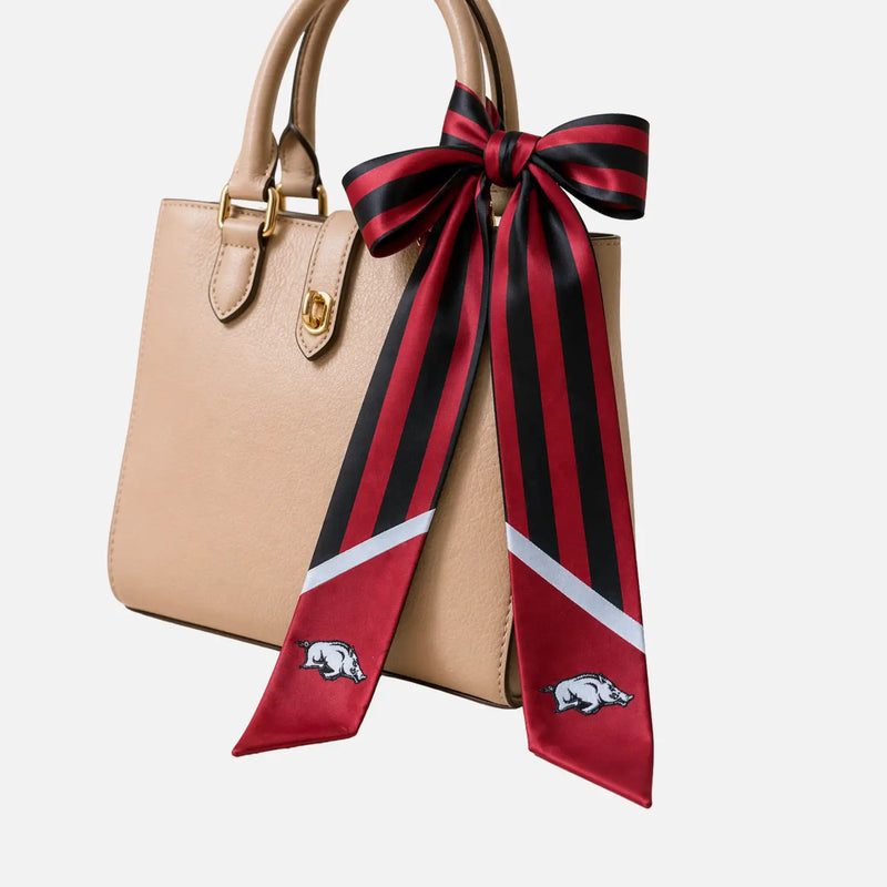 A beige leather handbag with gold-tone hardware and a large red, black, and white striped silk bow tied around the handle. The bow features the Arkansas Razorbacks logo near the bottom.