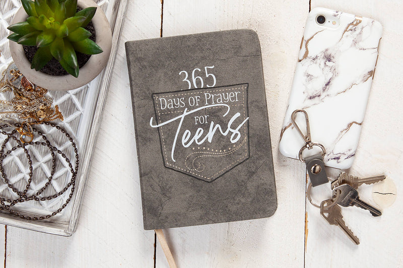 Gray prayer journal titled '365 Days of Prayer for Teens' on a wooden surface with a plant, phone, and keys.