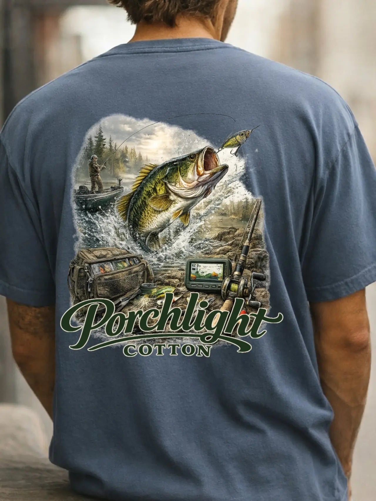 A man viewed from behind wearing a slate blue t-shirt with a large back graphic. The graphic features a bass jumping out of water, a fishing cabin, a fish finder, and the text 'Porchlight COTTON'.
