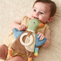 A duck-shaped baby teething toy with a green head, blue wings, brown body, orange beak and feet, a cream ring handle, and colorful fabric tabs along the tail, shown on a white background being held by a baby.