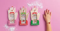 Three hand-shaped jewelry display boxes in pink, red, and green, each holding multiple small jewelry pieces, arranged on a pink background with scattered white powder. A human hand with painted nails and rings rests on the right side.
