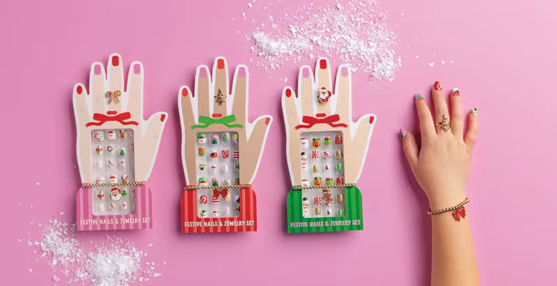 Three hand-shaped jewelry display boxes in pink, red, and green, each holding multiple small jewelry pieces, arranged on a pink background with scattered white powder. A human hand with painted nails and rings rests on the right side.