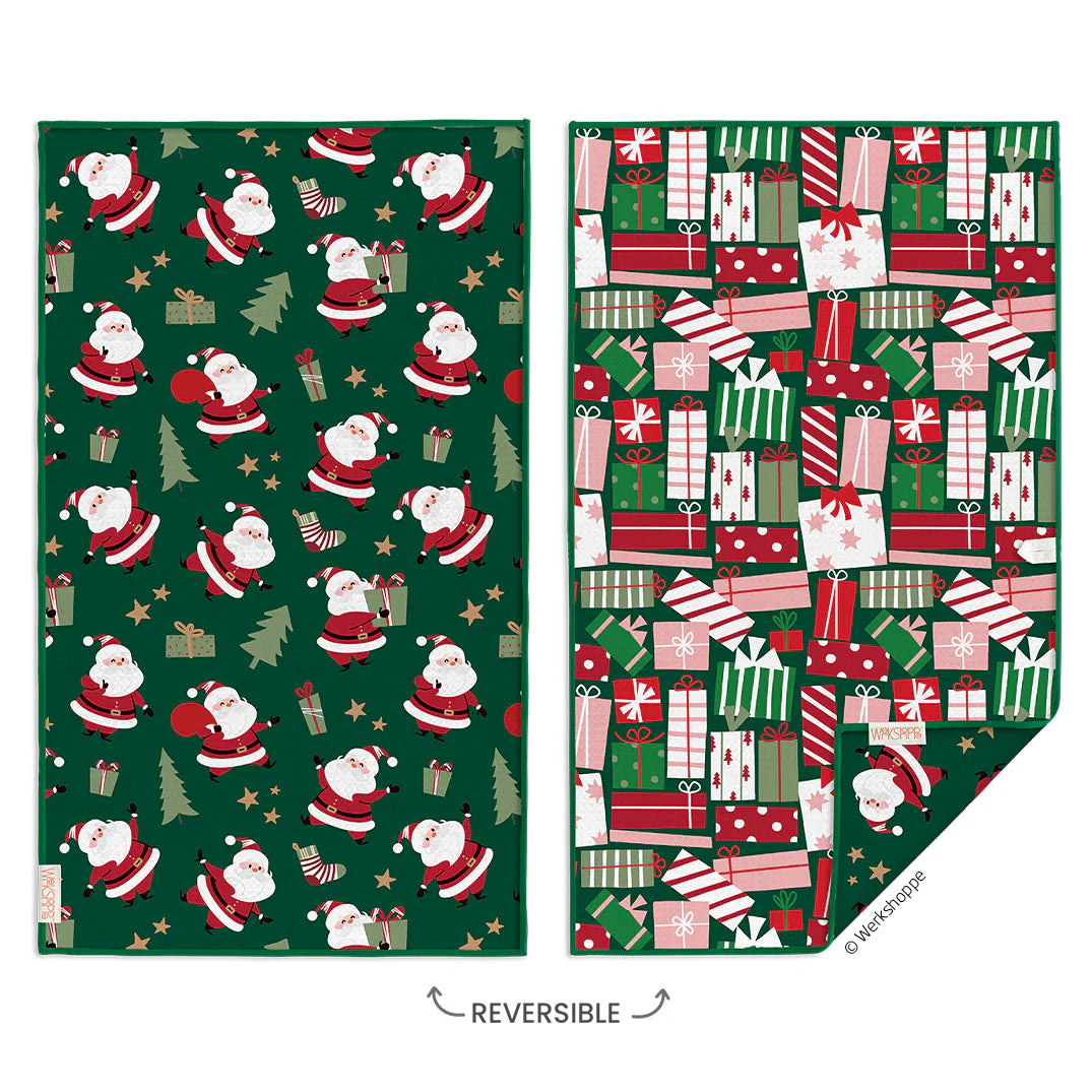 A reversible holiday-themed fabric or blanket shown in two views on a white background. The left side shows a dark green pattern with Santa Claus, Christmas trees, and gift boxes; the right side shows the reverse side with a dense pattern of wrapped presents in red, green, white, and pink.