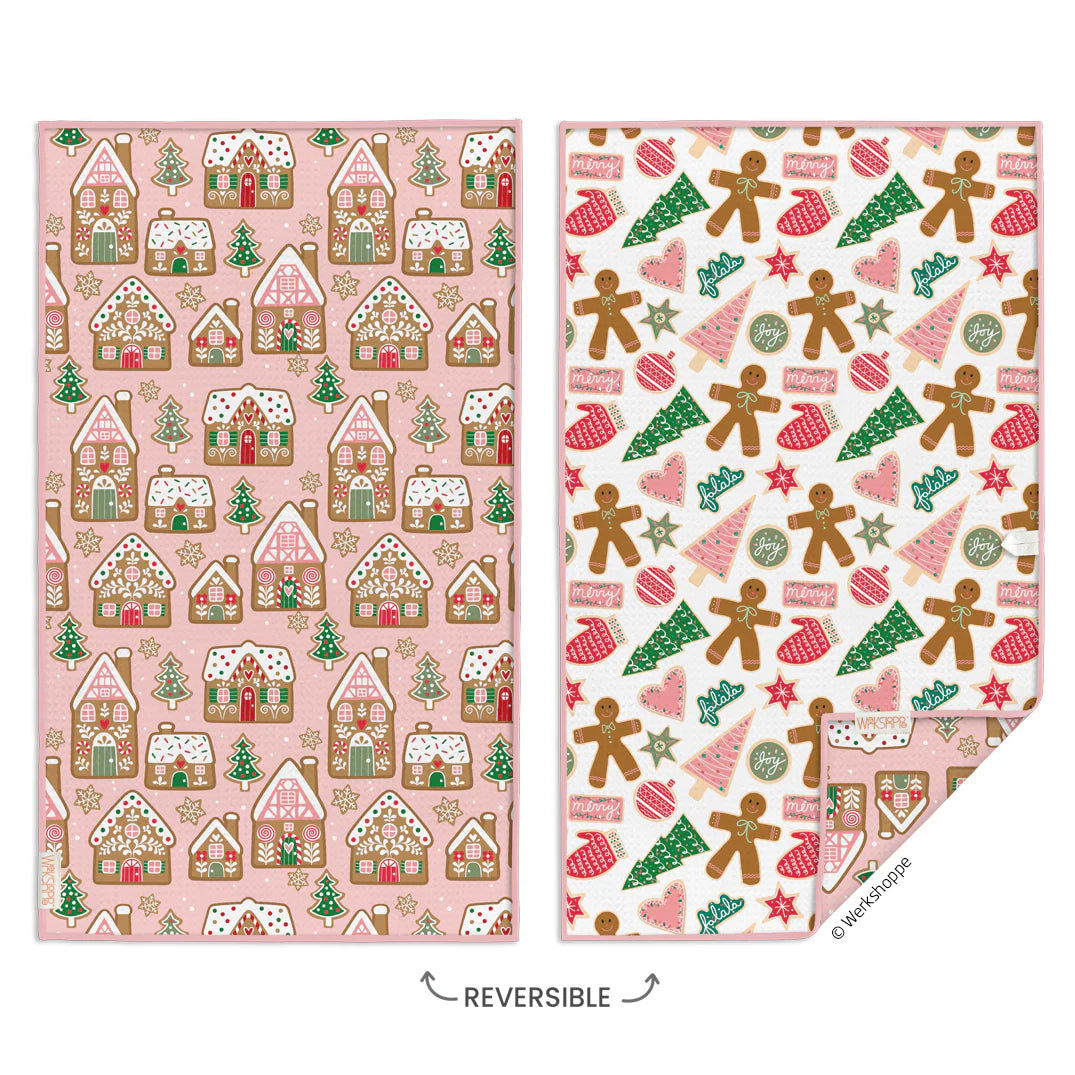 Two reversible holiday-themed fabric cloths shown side by side on a white background. The left cloth has a pink background with gingerbread houses, trees, and stars.