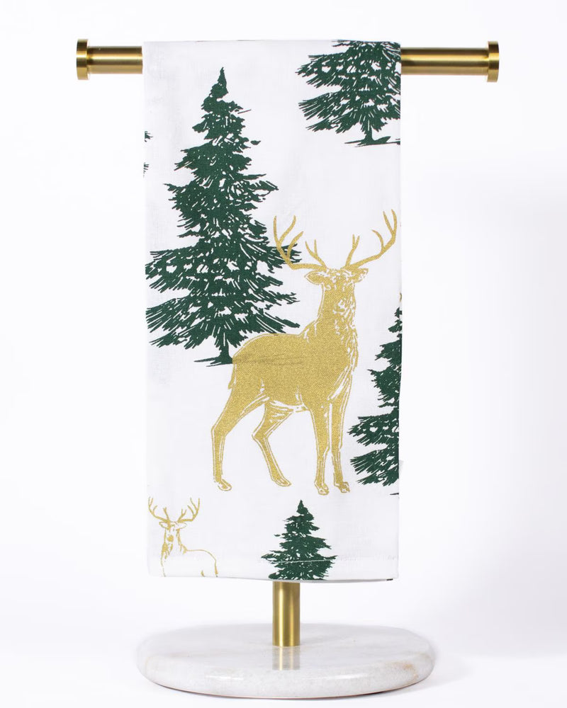 A white fabric hanging towel or scarf with a forest pattern featuring dark green pine trees and a large gold deer, displayed on a gold metal stand with a round marble base against a white background.
