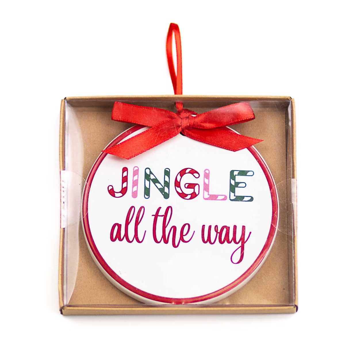 A round white Christmas ornament with a red ribbon loop and the phrase 'JINGLE all the way' in decorative lettering, presented inside a clear-front brown gift box on a white background.