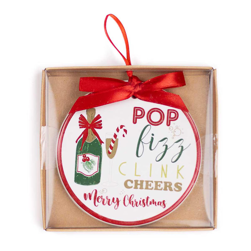 A round Christmas ornament in a clear square gift box with a kraft paper insert. The ornament is white with red and green text reading 'POP fizz CLINK CHEERS Merry Christmas' and features illustrations of a green champagne bottle, a candy cane, and a red ribbon bow.