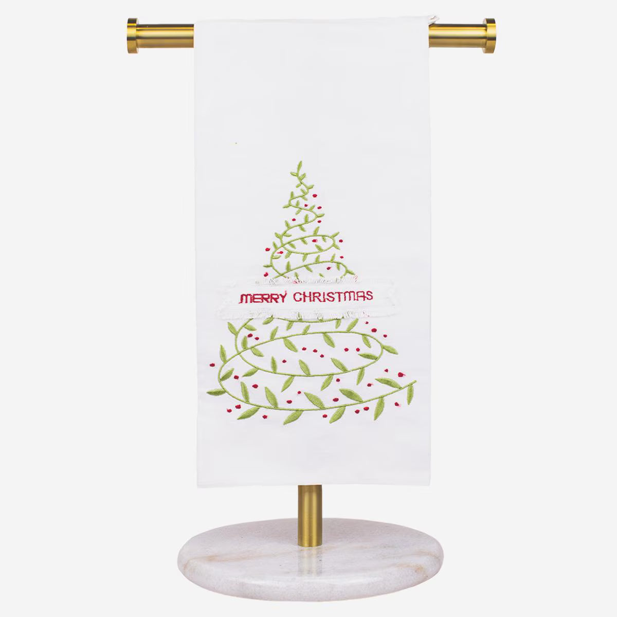 A white kitchen towel with a green and red Christmas tree design and the words 'MERRY CHRISTMAS' in red, hanging from a gold metal stand with a round white marble base against a light gray background.