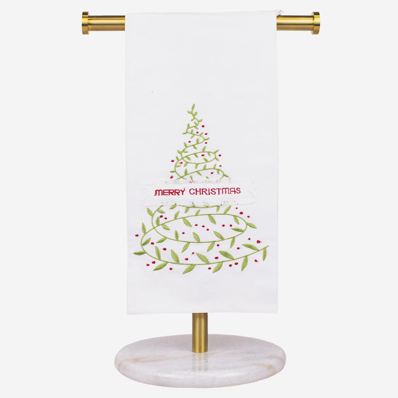 A white kitchen towel with a green and red Christmas tree design and the words 'MERRY CHRISTMAS' in red, hanging from a gold metal stand with a round white marble base against a light gray background.