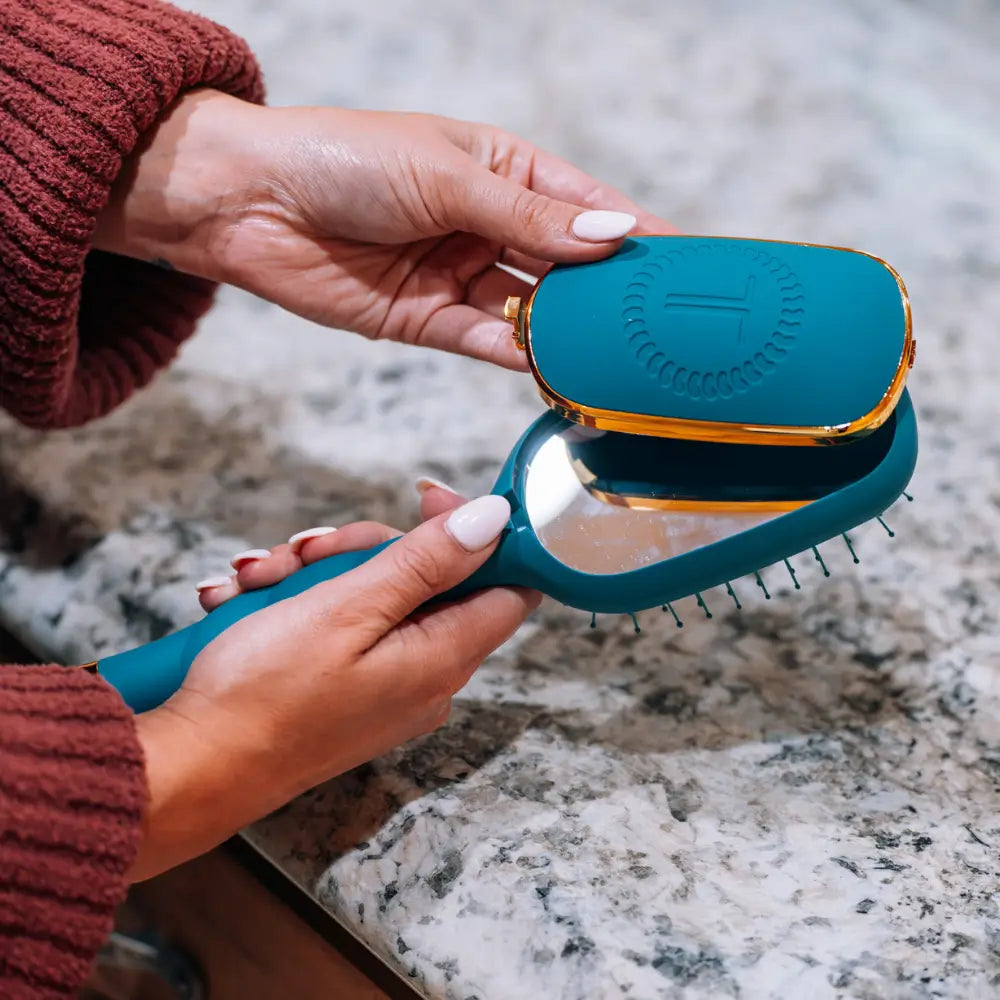 A teal and gold hairbrush with a built-in mirror, held by hands over a speckled granite countertop.