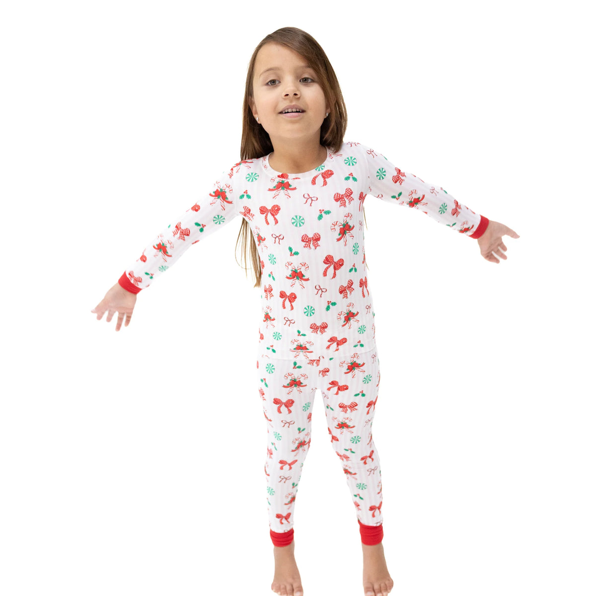 A young girl stands barefoot against a plain white background wearing a long-sleeve one-piece pajama top and pants set with a white base and a festive holiday print of red bows, green ornaments, and red cuffs.