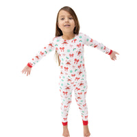 A young girl stands barefoot against a plain white background wearing a long-sleeve one-piece pajama top and pants set with a white base and a festive holiday print of red bows, green ornaments, and red cuffs.