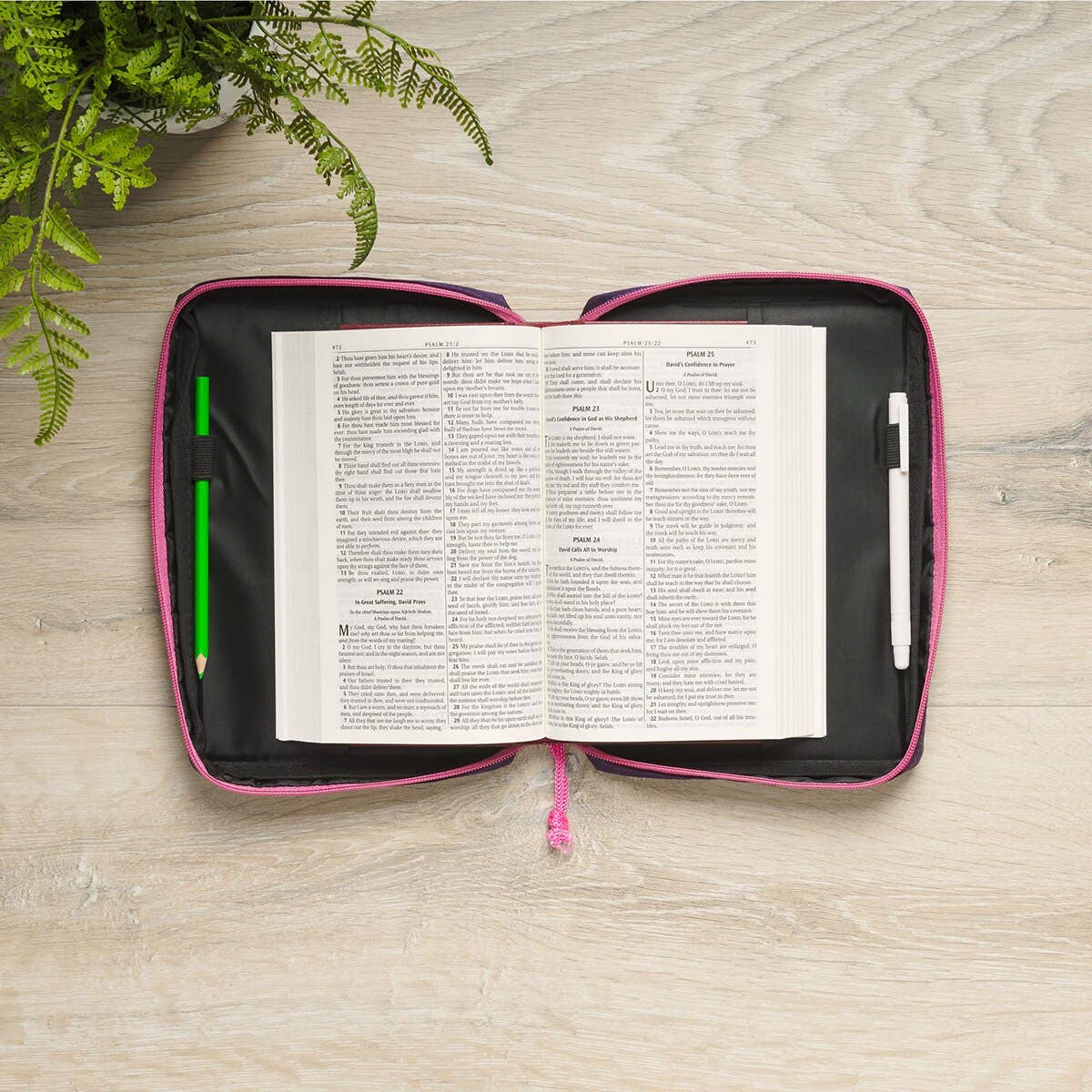 Open book in a pink and black zippered case on a wooden surface with greenery.