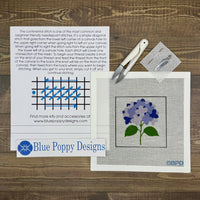 Cross-stitch kit with pattern, needle, thread, and Blue Poppy Designs branding on a wooden surface.