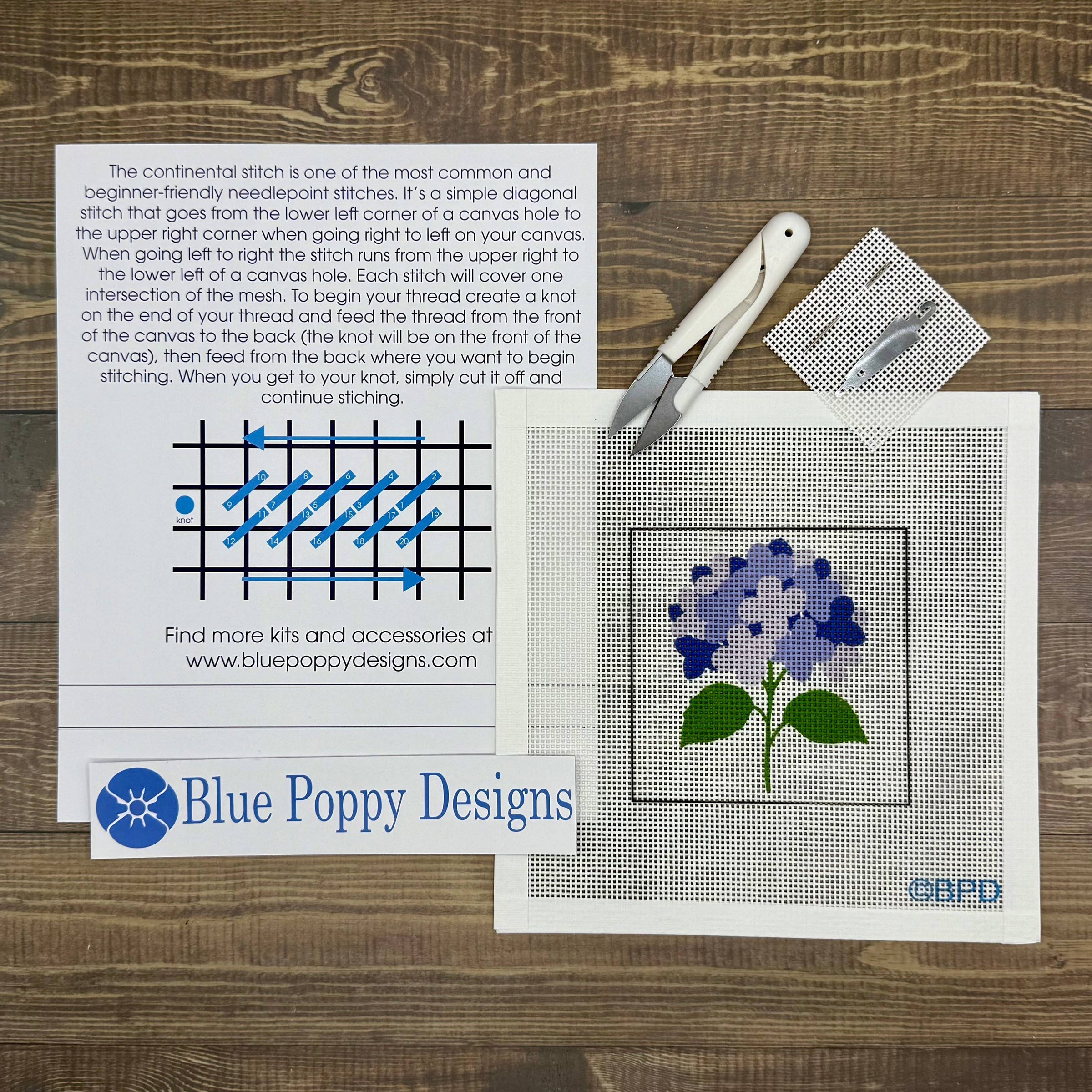 Cross-stitch kit with pattern, needle, thread, and Blue Poppy Designs branding on a wooden surface.