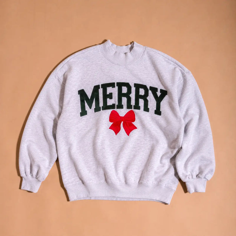 A light heather gray crewneck sweatshirt featuring the word 'MERRY' in dark green varsity-style letters with a red bow graphic underneath, laid flat on a warm tan background.