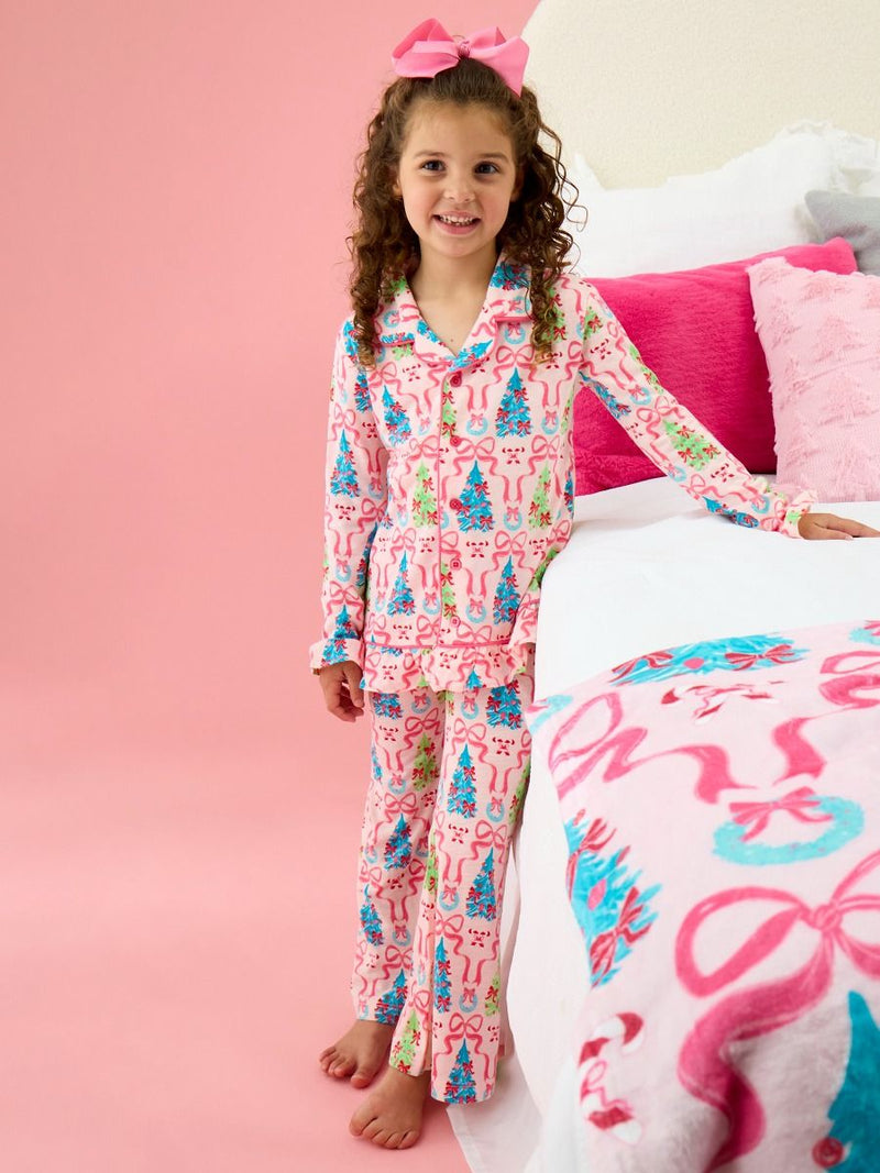 A young girl with curly brown hair wearing a pink pajama set with a Christmas tree and candy cane pattern, standing next to a bed with pink bedding.