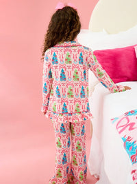 A child wearing a pink long-sleeve pajama set with a colorful Christmas tree and bow pattern, shown from the back in a bedroom setting with a pink wall and bed.