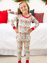 A young girl stands in front of a bed wearing a festive Christmas-themed long-sleeve pajama set. The pajamas feature a light cream background with a repeating pattern of red bows, green holly leaves, and pinecones, with red trim on the collar, cuffs, and ankles.