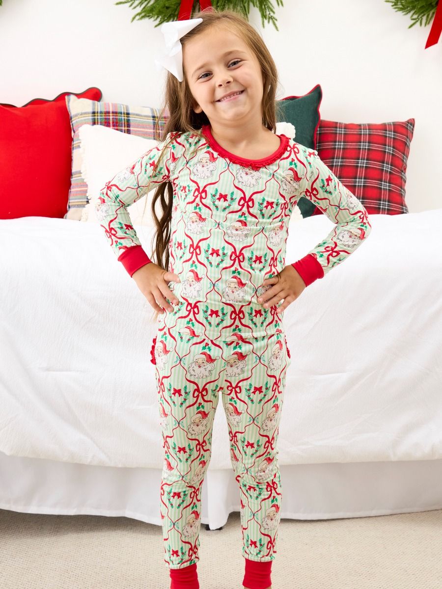 A young girl stands in front of a bed wearing a festive Christmas-themed long-sleeve pajama set. The pajamas feature a light cream background with a repeating pattern of red bows, green holly leaves, and pinecones, with red trim on the collar, cuffs, and ankles.