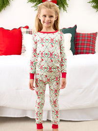 A young girl stands in front of a white bed with holiday-themed pillows, wearing a long-sleeve Christmas-themed pajama set with a red and green patterned design.