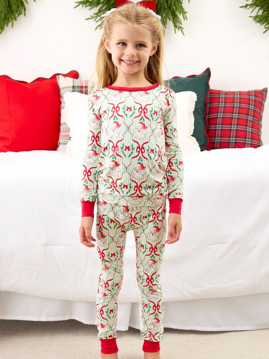 A young girl stands in front of a white bed with holiday-themed pillows, wearing a long-sleeve Christmas-themed pajama set with a red and green patterned design.