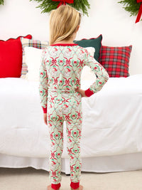 A child standing with their back to the camera wearing a long-sleeve Christmas-themed pajama set with a red and green holiday pattern on a white background, posed in front of a bed with holiday pillows.
