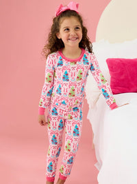 A young girl with curly brown hair wearing a pink long-sleeve pajama set with a Christmas tree and candy cane pattern, standing next to a white bed with a pink pillow against a pink background.