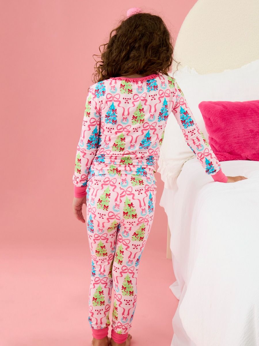 A child wearing a pink long-sleeve pajama set with a colorful Christmas tree and bow pattern, shown from the back in a bedroom setting with a pink wall and white bed.