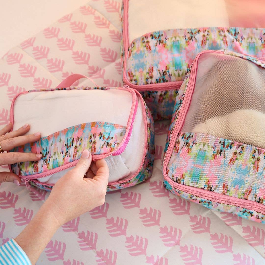 Set of floral-patterned travel bags with a hand interacting with one of them on a pink leaf-patterned surface.