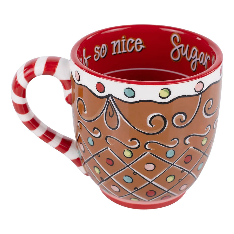 A festive ceramic mug designed to look like a gingerbread cookie, featuring a brown body with white icing-like borders, colorful candy dots, swirl patterns, a red interior with white script text, and a red-and-white striped handle shaped like a candy cane.