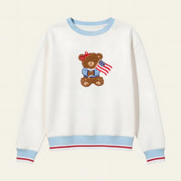 White sweatshirt with a teddy bear design holding an American flag on a beige background