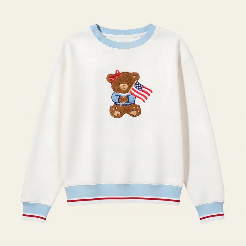 White sweatshirt with a teddy bear design holding an American flag on a beige background