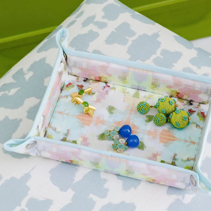 A rectangular fabric jewelry organizer tray with a pastel floral print and light blue trim, shown holding several pairs of earrings. The tray is placed on a white surface with a gray abstract pattern and a bright green background visible in the upper left.