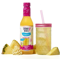 Skinny Mixes Pineapple Whip drink bottle with a pink cap, pineapple slices, and a glass with pink lid and straw on a white background.