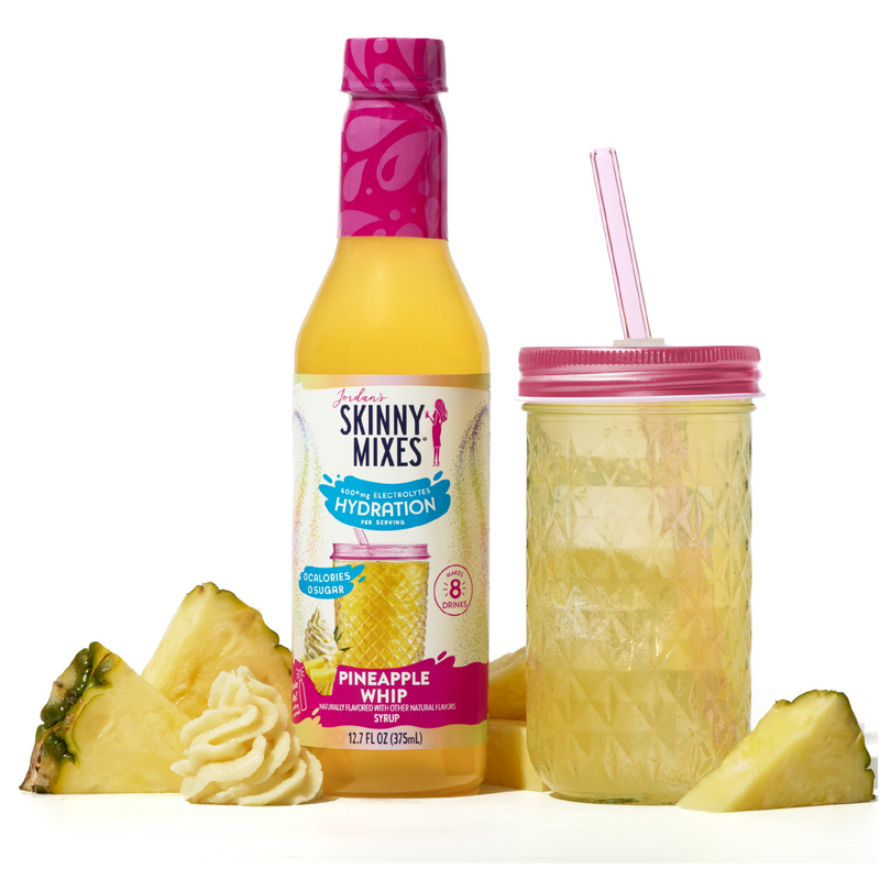 Skinny Mixes Pineapple Whip drink bottle with a pink cap, pineapple slices, and a glass with pink lid and straw on a white background.