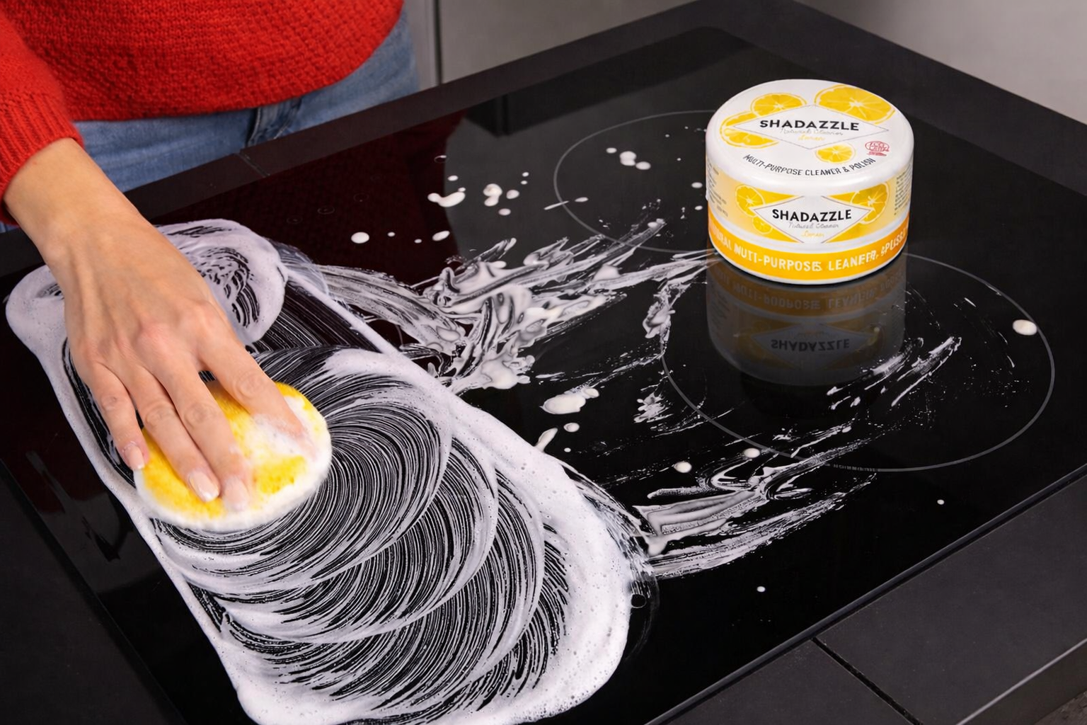Person cleaning a glass surface with a sponge and Shadazze cleaning product.