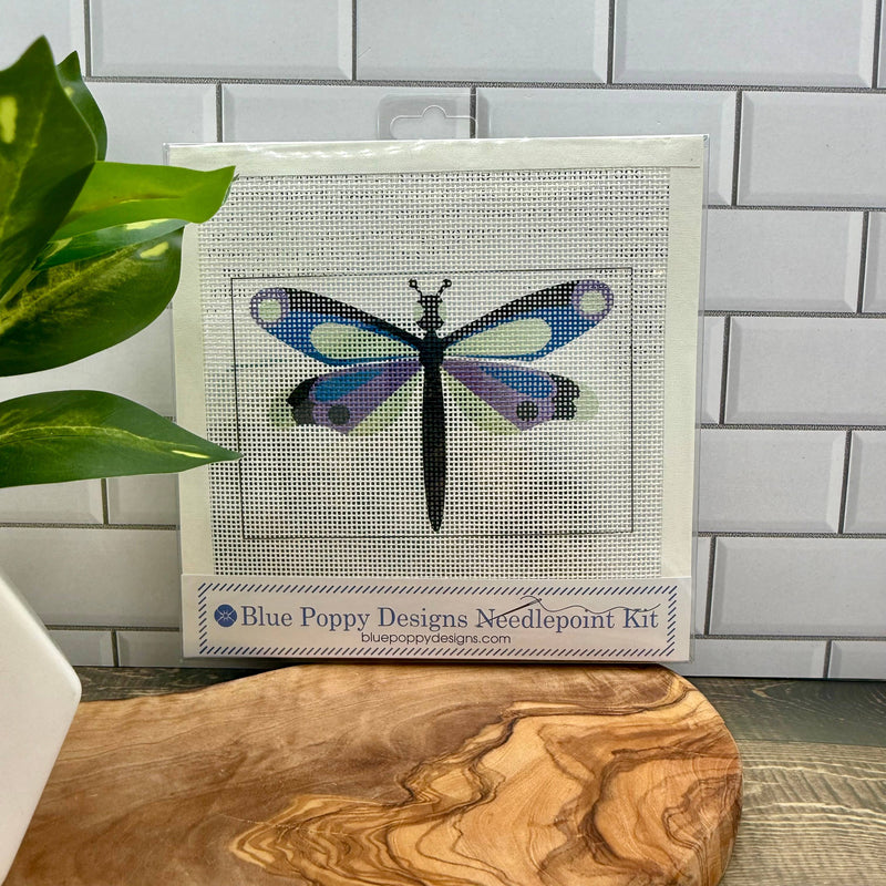 Blue Poppy Designs Needlepoint Kit with a dragonfly design on a wooden surface against a tiled wall.
