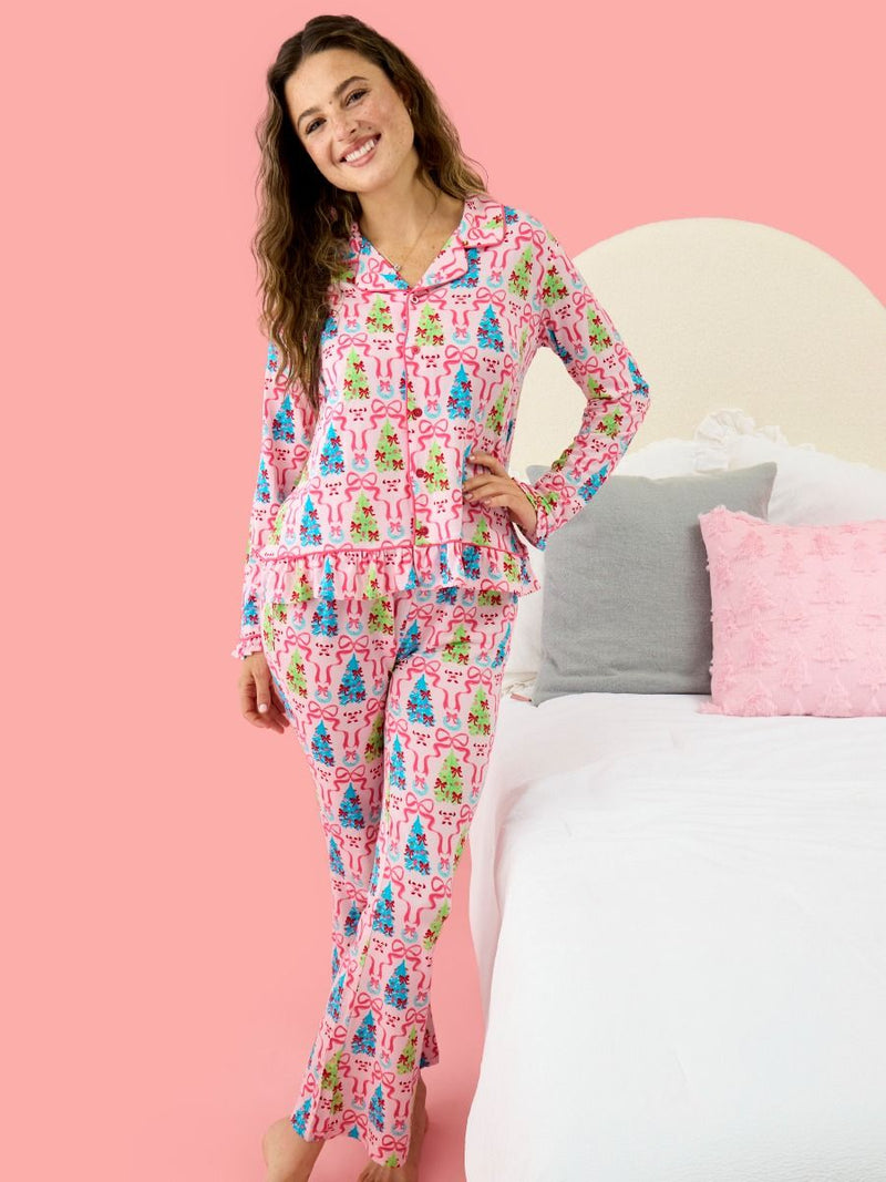 A woman modeling a pink long-sleeve pajama set with a colorful Christmas tree pattern, standing in front of a pink wall and a bed with white bedding and pillows.