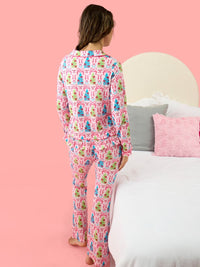 A woman stands with her back to the camera wearing a pink long-sleeve pajama set with a colorful Christmas tree pattern, positioned next to a bed with white and pink bedding against a solid pink wall.