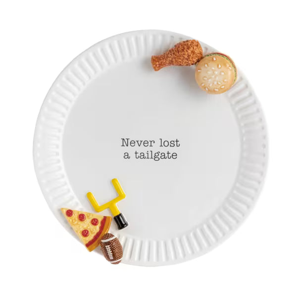 A white ceramic platter with a black printed phrase reading 'Never lost a tailgate' in the center, styled with small food-themed decorations including a chicken leg, hamburger bun, pizza slice, football, and a yellow goal post on a white background.