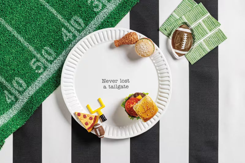 A white ceramic platter with the phrase 'Never lost a tailgate' in black text, styled with football-themed props on a black and white striped surface. The plate includes a small chicken nugget, a biscuit, a hamburger, a pizza slice, and a toy lightning bolt.