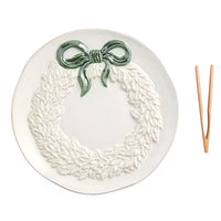 A white ceramic plate featuring a raised relief wreath design with a glossy green bow at the top, shown on a clean white background with a pair of wooden serving utensils to the right.