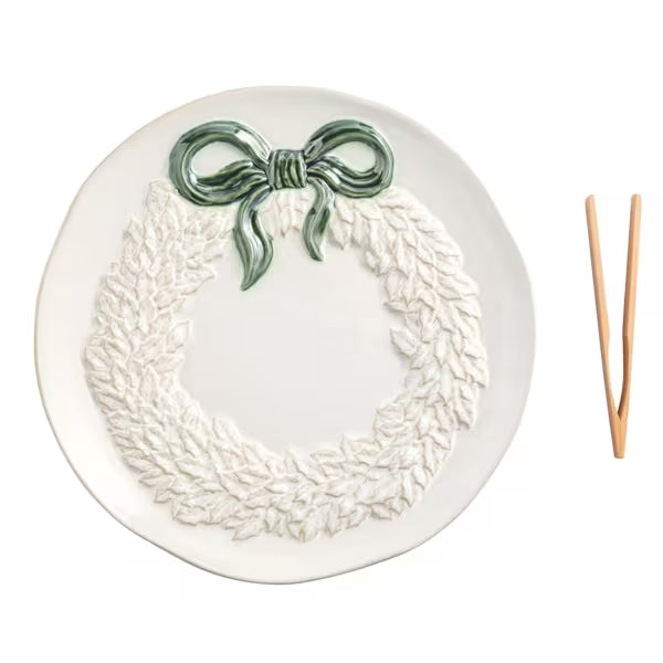 A white ceramic plate featuring a raised relief wreath design with a glossy green bow at the top, shown on a clean white background with a pair of wooden serving utensils to the right.