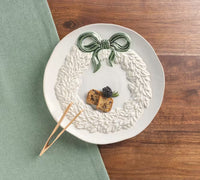 A white ceramic plate with a raised leaf wreath design and a green bow, styled on a wooden table with a green napkin and wooden picks.