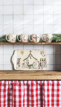 A rustic ceramic Christmas nativity scene serving platter displayed on a wooden shelf in a kitchen setting, with a red and white gingham checkered curtain below and other decorated mugs on the shelf above.