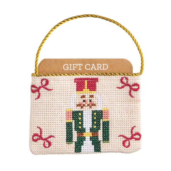 A small cross-stitch gift card holder featuring a nutcracker design and pink bows on a cream fabric background, with a gold rope handle and a brown backing card labeled 'GIFT CARD'.