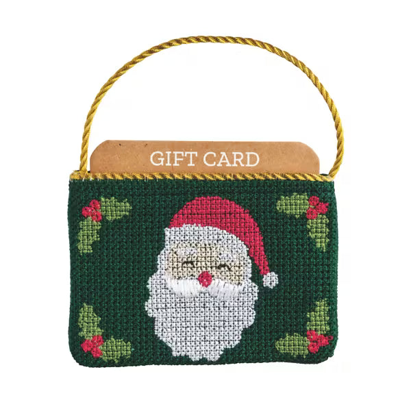 A Christmas-themed needlepoint canvas bag with a gold rope handle, featuring a stitched Santa Claus face on a dark green background with holly accents. A brown gift card is tucked inside the top opening, which is labeled 'GIFT CARD'.