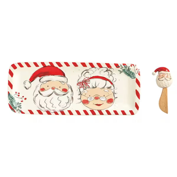 A rectangular Christmas-themed serving tray with a red and white striped border, featuring illustrated Santa Claus and an elderly woman with holiday decorations. A small wooden spoon with a Santa head topper is shown to the right on a plain white background.