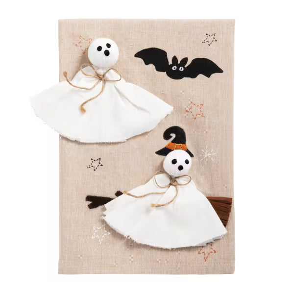 A beige fabric tea towel or hanging textile featuring Halloween-themed appliques, including two white ghost figures with black embroidered faces, a black bat, orange star accents, and a witch figure with a black hat and broom.